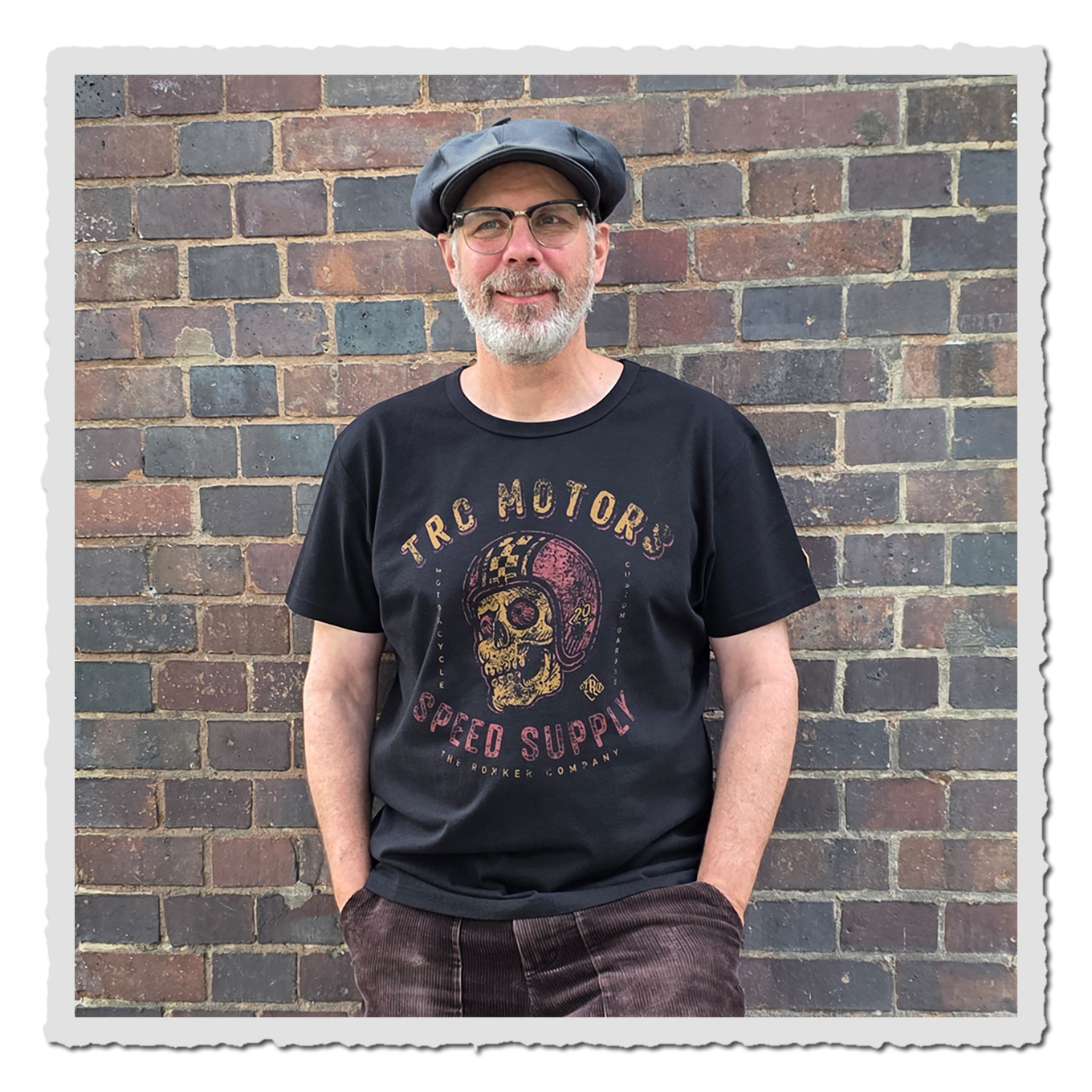 Person trägt schwarzes T-Shirt mit mehrfarbigem Skull-Print vor einer braun-grauen Backsteinwand.